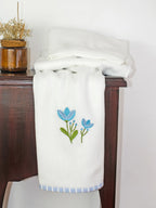 White Kerala bath towel with blue floral embroidery and a blue striped border on a wooden chest