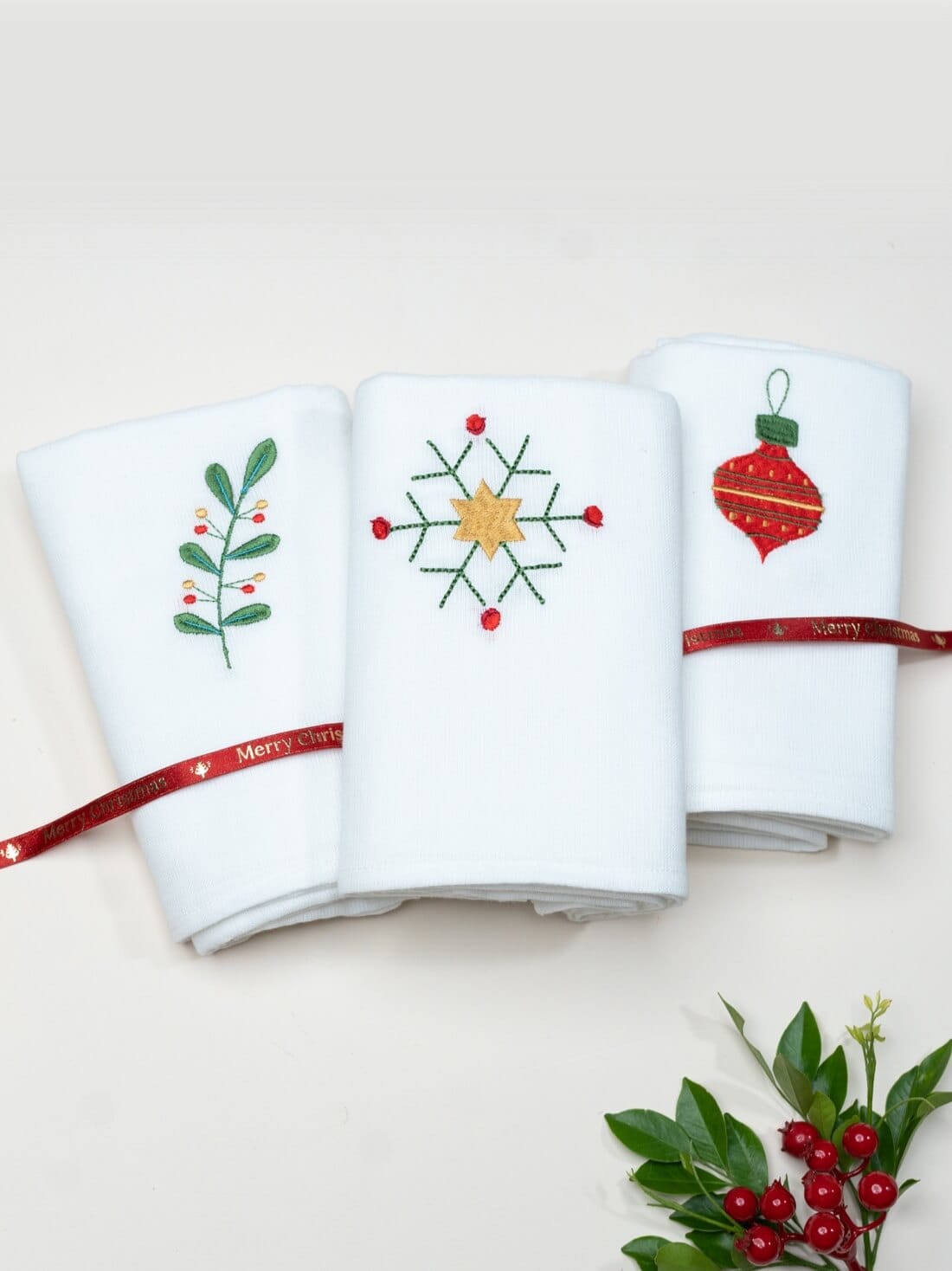 Three folded white towels embroidered with Christmas berries, a star snowflake and a red ornament