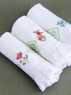 Three folded white Kerala bath towels with pink, red and blue embroidered motifs and ruffled edges