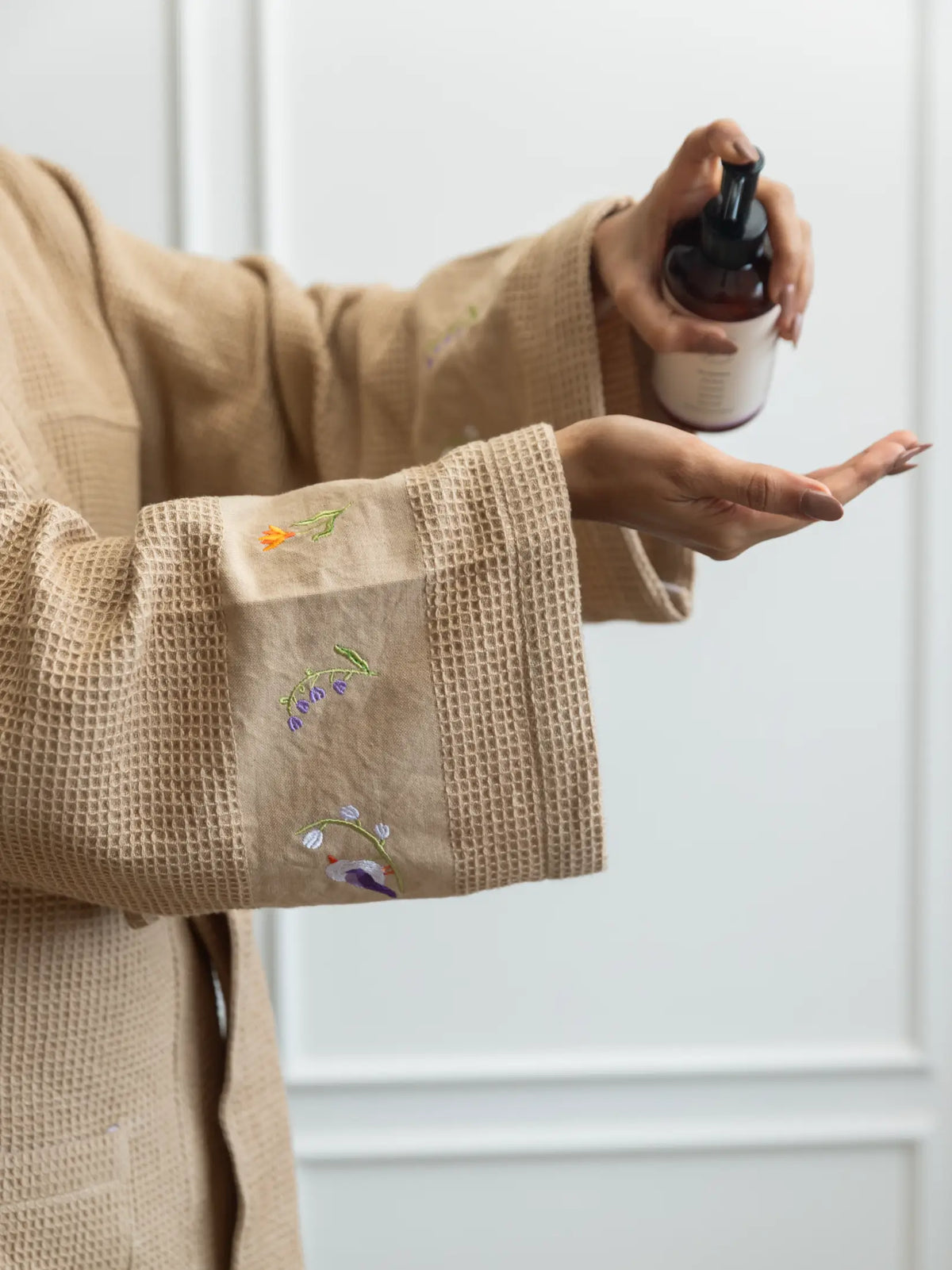 Woman applying lotion while wearing a beige waffle bath robe with embroidered cuffs