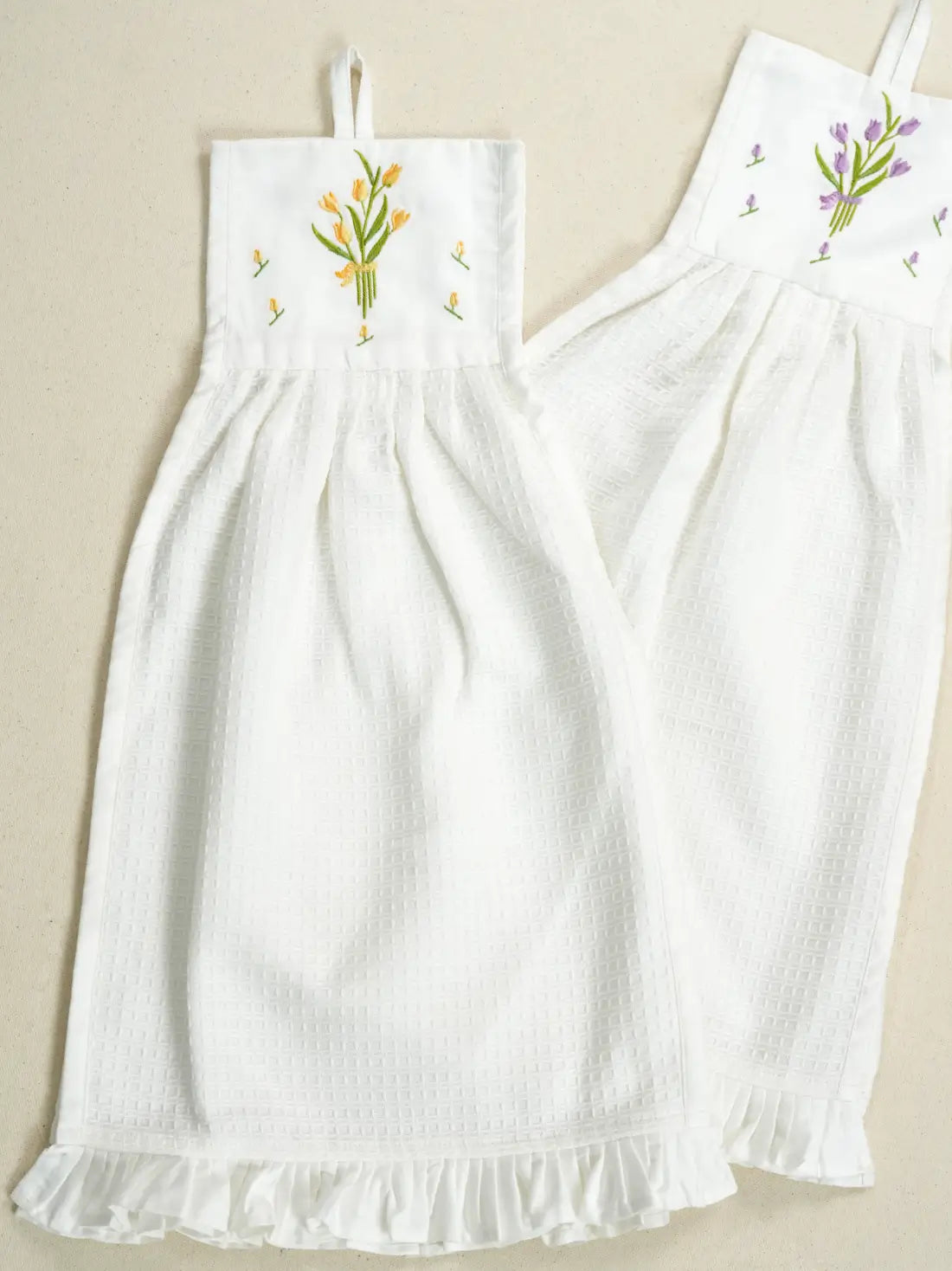 Two white hanging waffle hand towels with yellow and purple tulip embroidery and ruffled hems